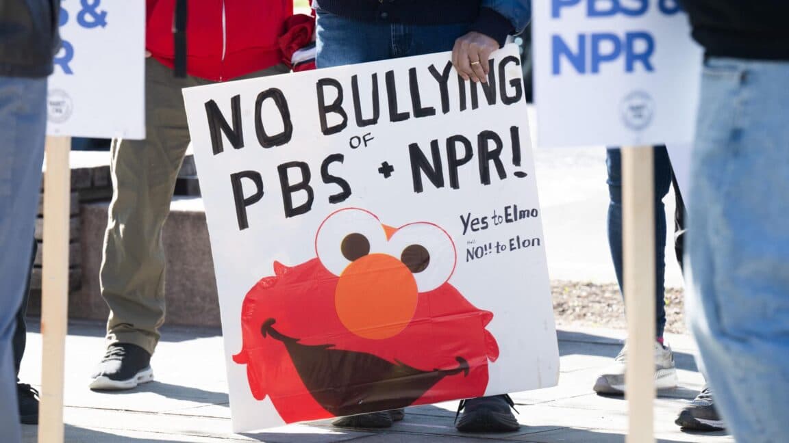 People at a rally urge Congress to protect funding for public broadcasters. A man carries a sign that has a picture of Sesame Street character Elmo and says, "No bullying of PBS and NPR: Yes to Elmo, No to Elon." Another sign says, "Defend PBS and NPR."
