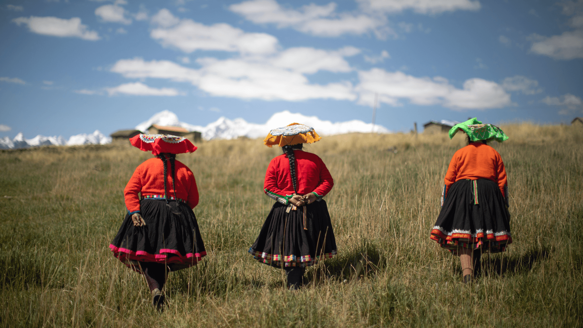 three women wearing red shirts and hats walk through a grassy field towards a mountain