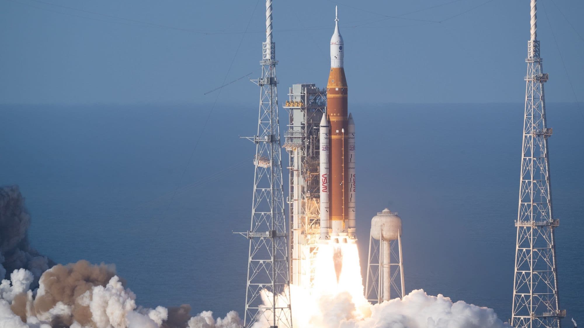 a huge orange and white rocket launches into a blue sky from a seaside pad