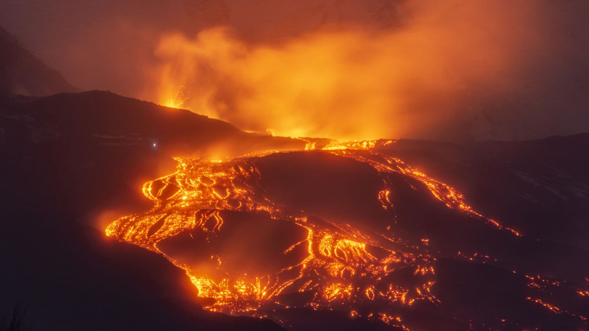 A volcano glowing with orange and yellow lava is seen against the evening night sky.