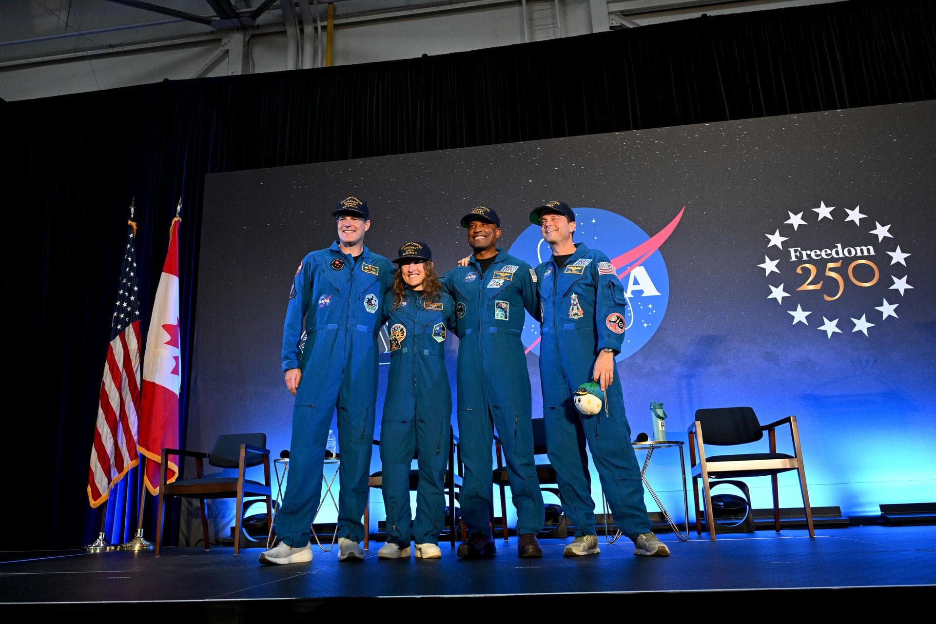 The four Artemis II crew members stand side-by-side and smiling in their blue flight suits on a stage at NASA Johnson. Behind them are the American and Canadian flags, NASA logo, and Freedom 250 logo.