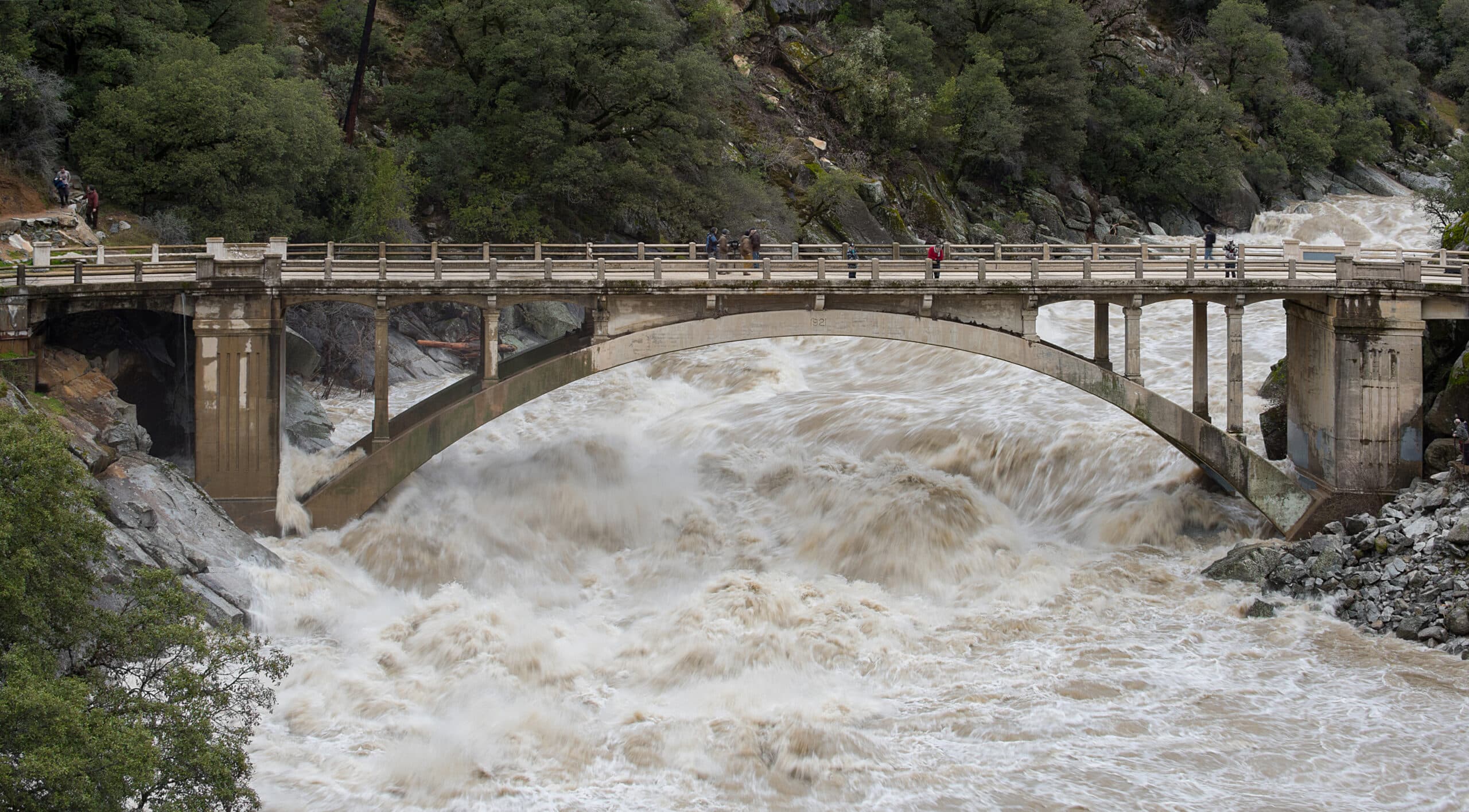 A photograph of a bridge suspended between two rocky hills. Underneath, foamy, brown water rages downstream. A family sits on the bridge watching the water.  (via news.agu.org)