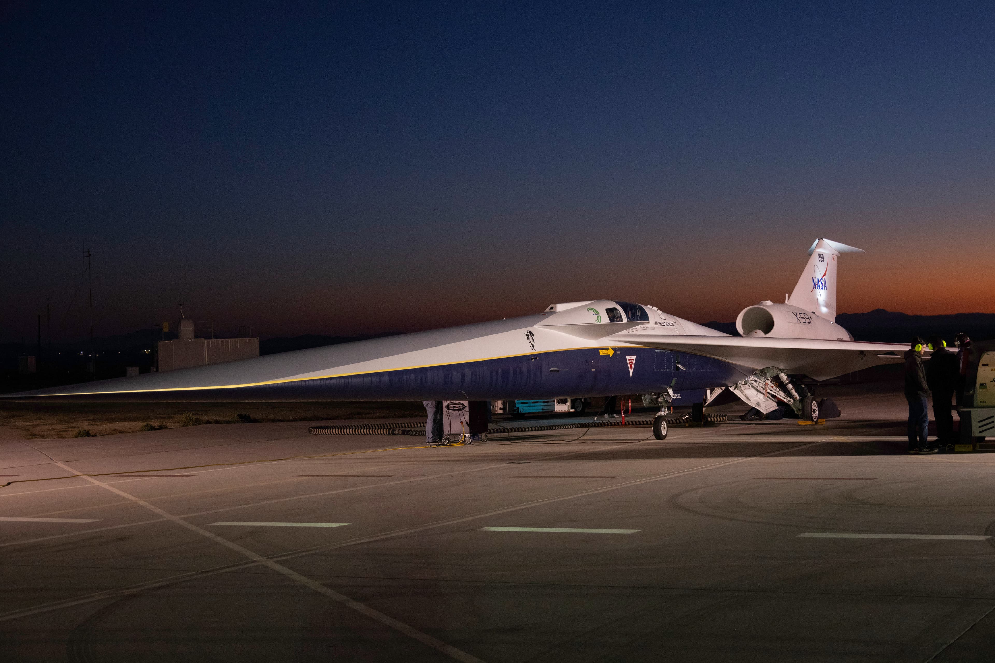 An aircraft resting on a section of runway as seen from the side. The X-59 has a long, thin nose that accounts for nearly a third of its length, along with sleek wings and an engine mounted above its body, just below its tail. The early morning sky is dark in the background with the sunrise just starting to emerge.