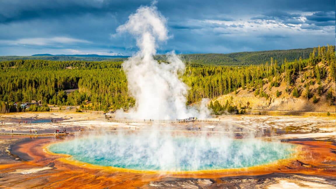Image of a circular pool of pale blue water with steam rising from it. The surroundings are richly covered soils with forests in the distance.