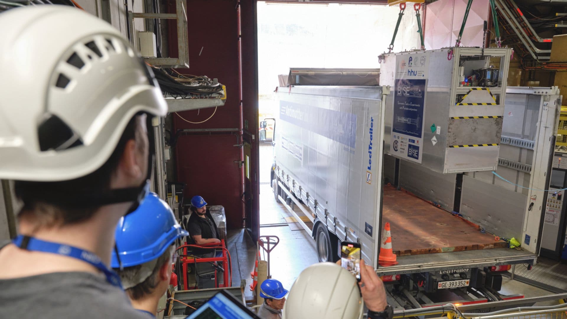 A view of a large white truck being loaded by crane with a large metal box. Workers wearing hard hats stand to the left of the truck.
