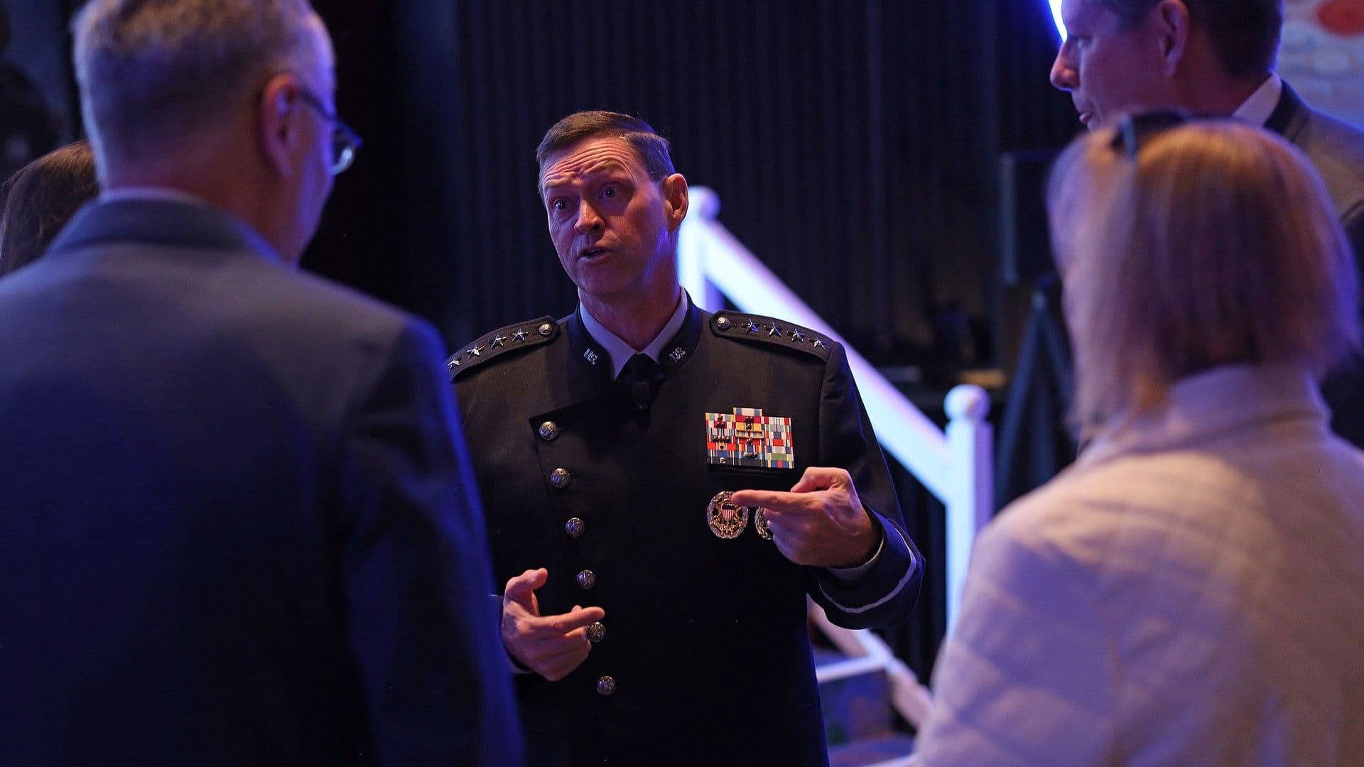 a man in a black military uniform speaks to a group of attendees on a conference ballroom floor