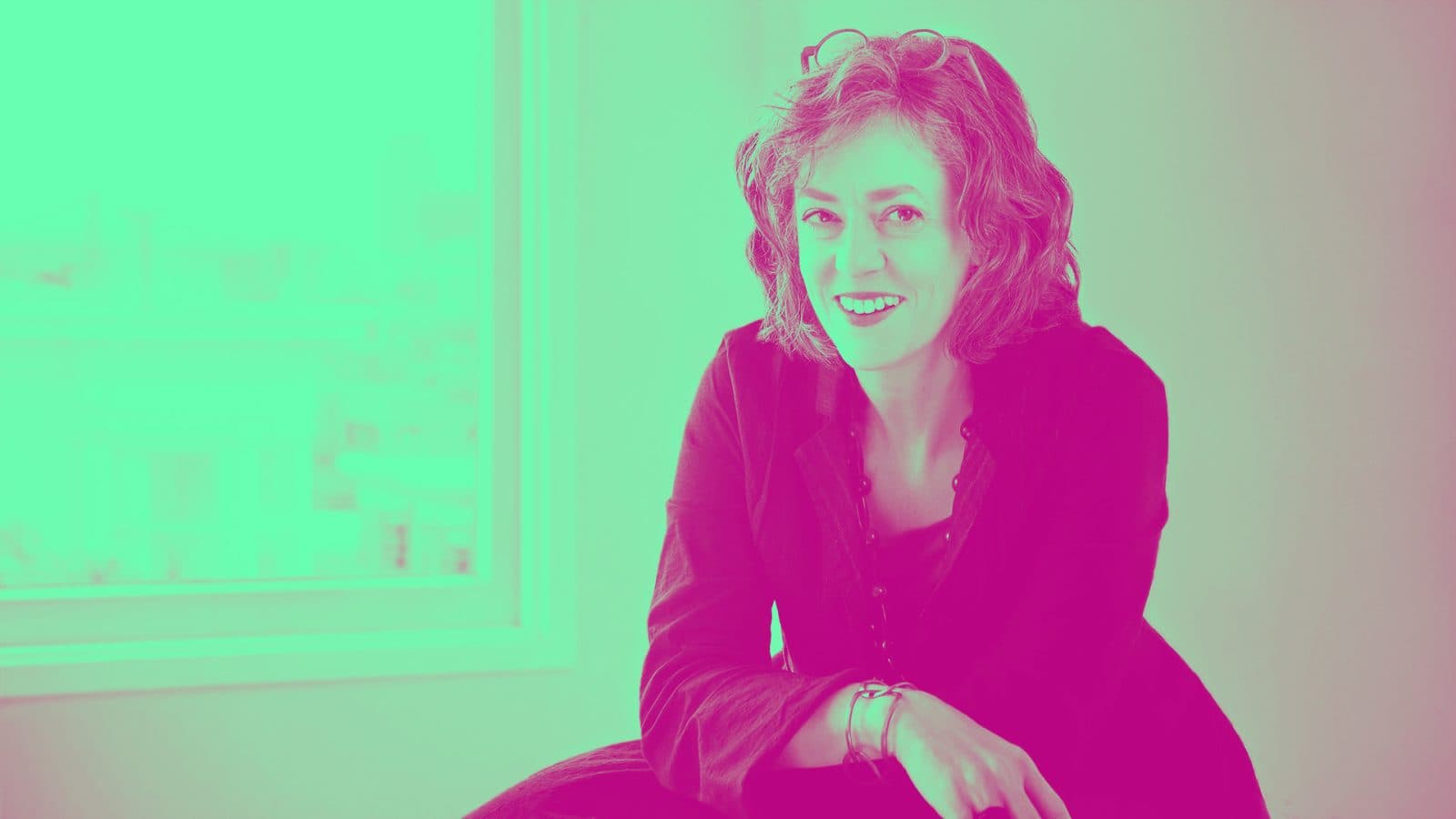 A smiling woman with curly hair sitting near a window, wearing a black jacket and bracelets, in a vibrant, pink-tinted setting.