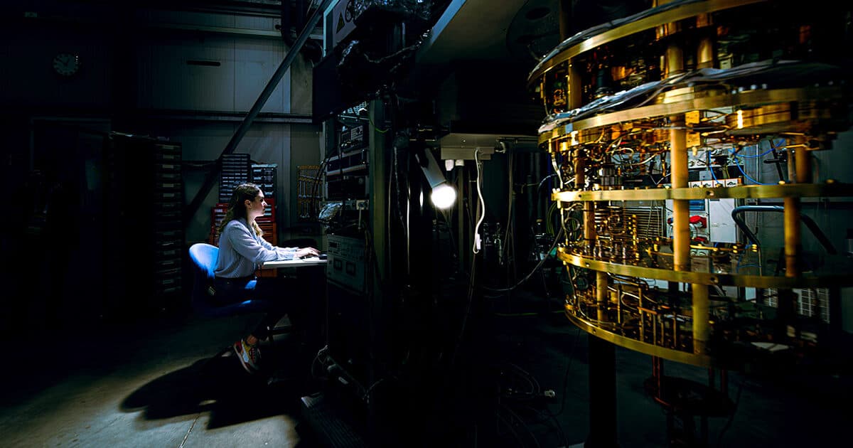 A researcher sits at a keyboard at a workstation illuminated by a single light in a darkened lab facility. To the right is a large piece of complex machinery featuring stacked rings of gold-colored metal components and intricate circuitry.