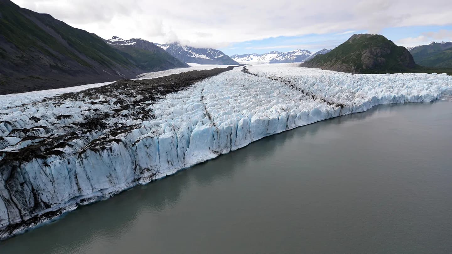 An aerial view shows a wide glacier flowing between mountains and ending at a gray glacial lake. (via sciencenews.org)